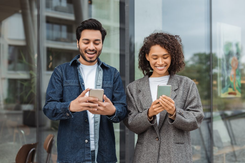 Two cheerful young professionals using smartphones outside a modern office, representing mobile-focused communication in SMS marketing Malaysia.