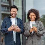 Two cheerful young professionals using smartphones outside a modern office, representing mobile-focused communication in SMS marketing Malaysia.