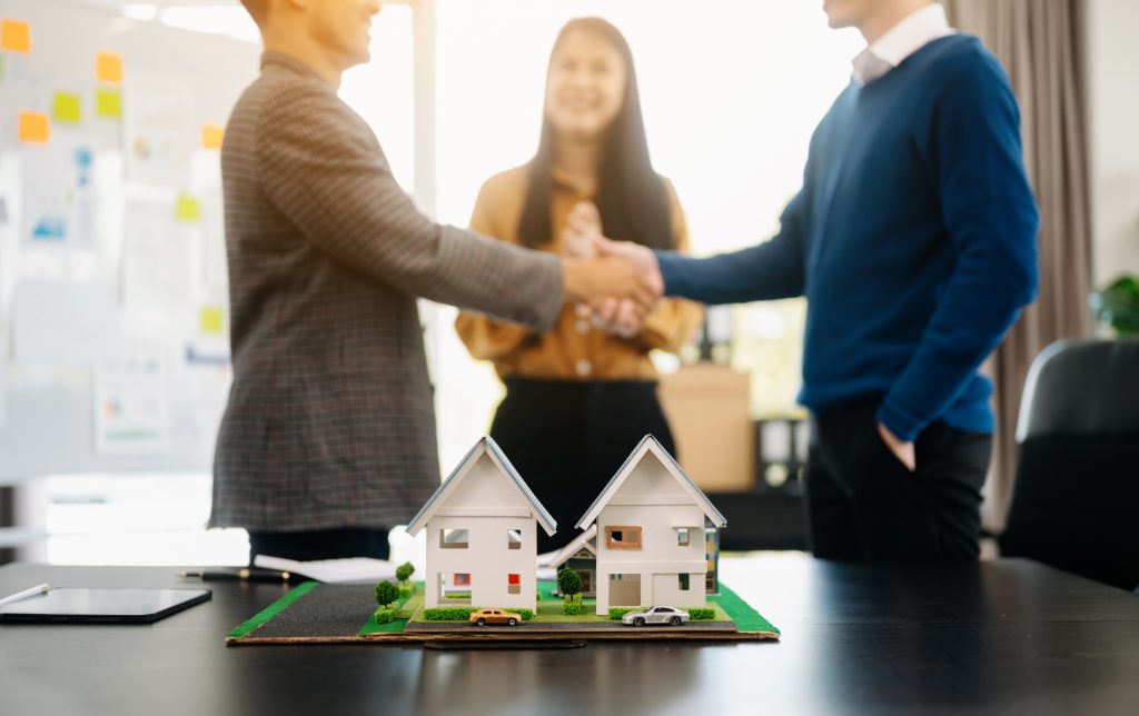 Two business professionals shaking hands in front of a smiling client, with a miniature house model on a desk, symbolizing a real estate deal — representing industries that benefit from the best SMS service provider.