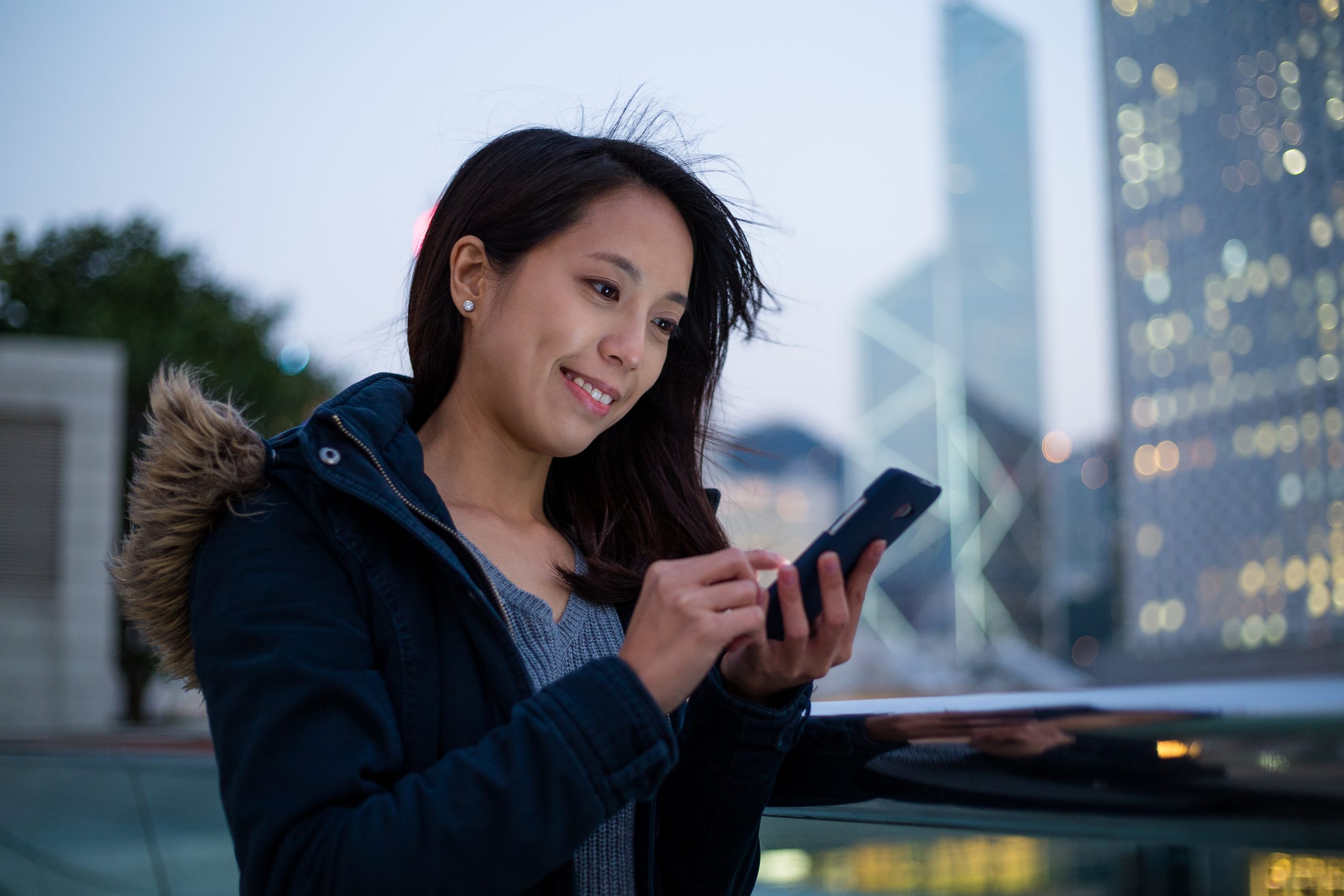 Young woman using a smartphone in a city at dusk, representing secure customer messaging and SMS service Malaysia for businesses.