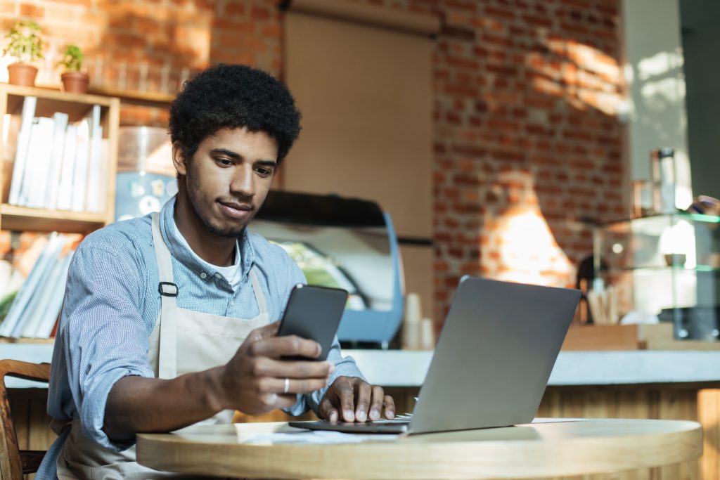 Small business owner in a café checks an SMS on his phone while using a laptop — illustrating SMS marketing Malaysia for fast customer engagement.