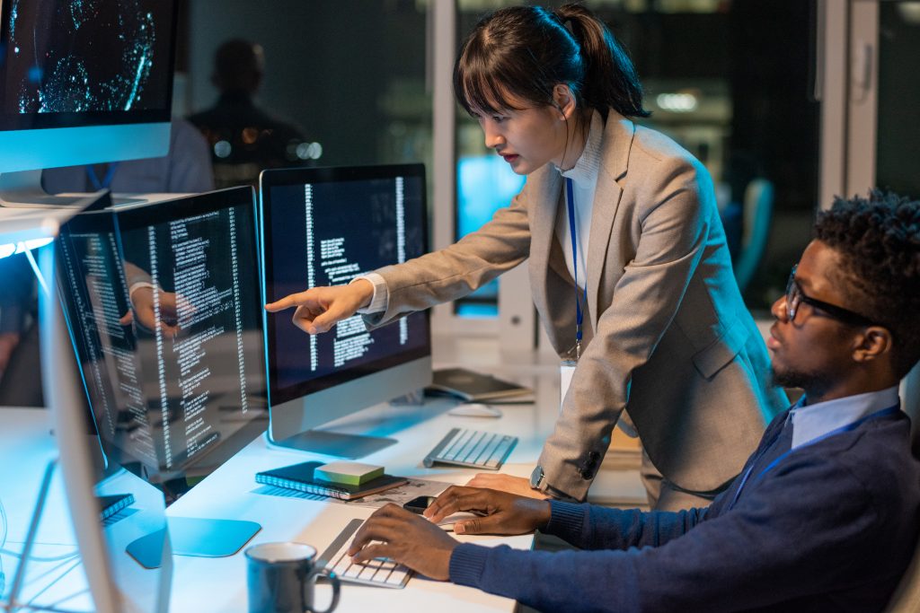 Female engineer explaining send SMS API code to colleague on computer screens in a modern office.