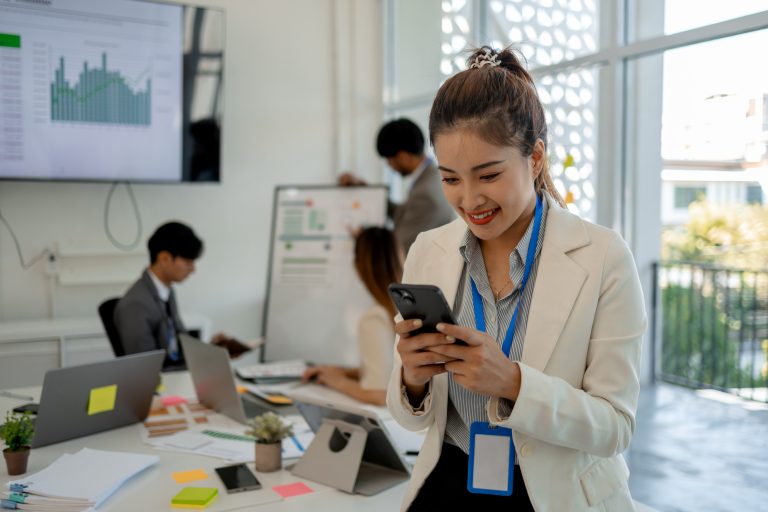 A businesswoman smiling while checking her phone in a modern office, representing global messaging success with the Best SMS Service Provider.