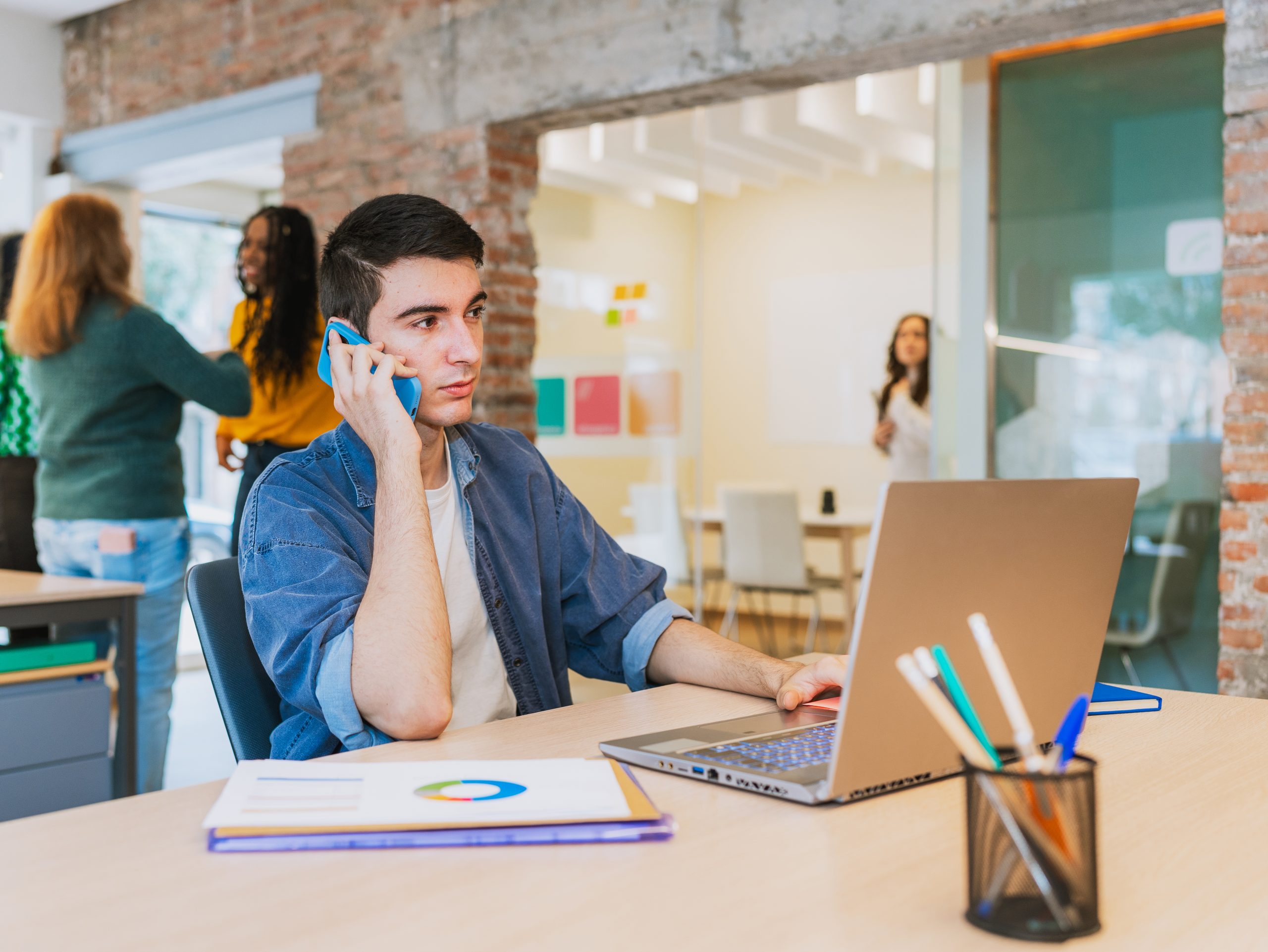 Young professional on phone call while working on laptop in a modern office, planning an SMS Marketing Malaysia campaign.
