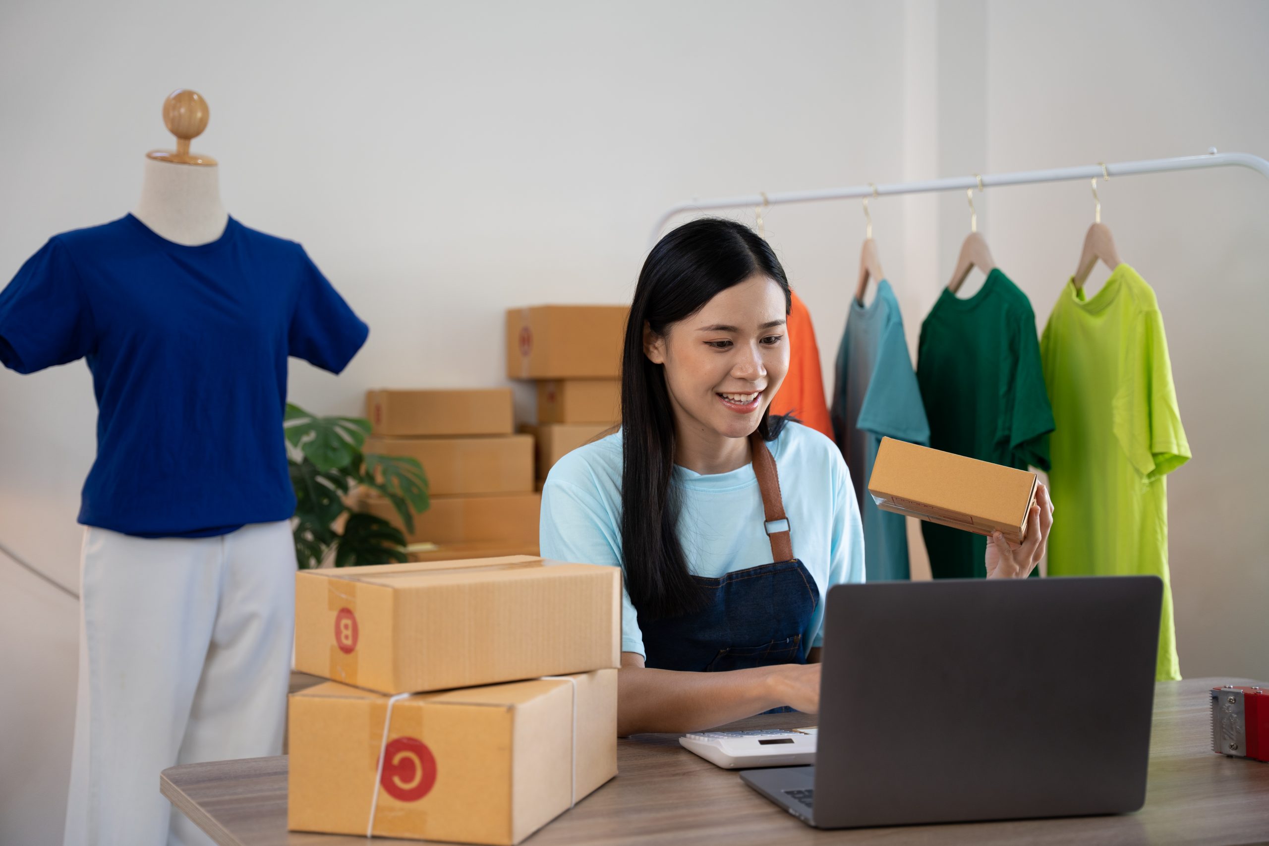 A young e-commerce owner smiling while packing products and managing orders on her laptop using an SMS Gateway Provider.