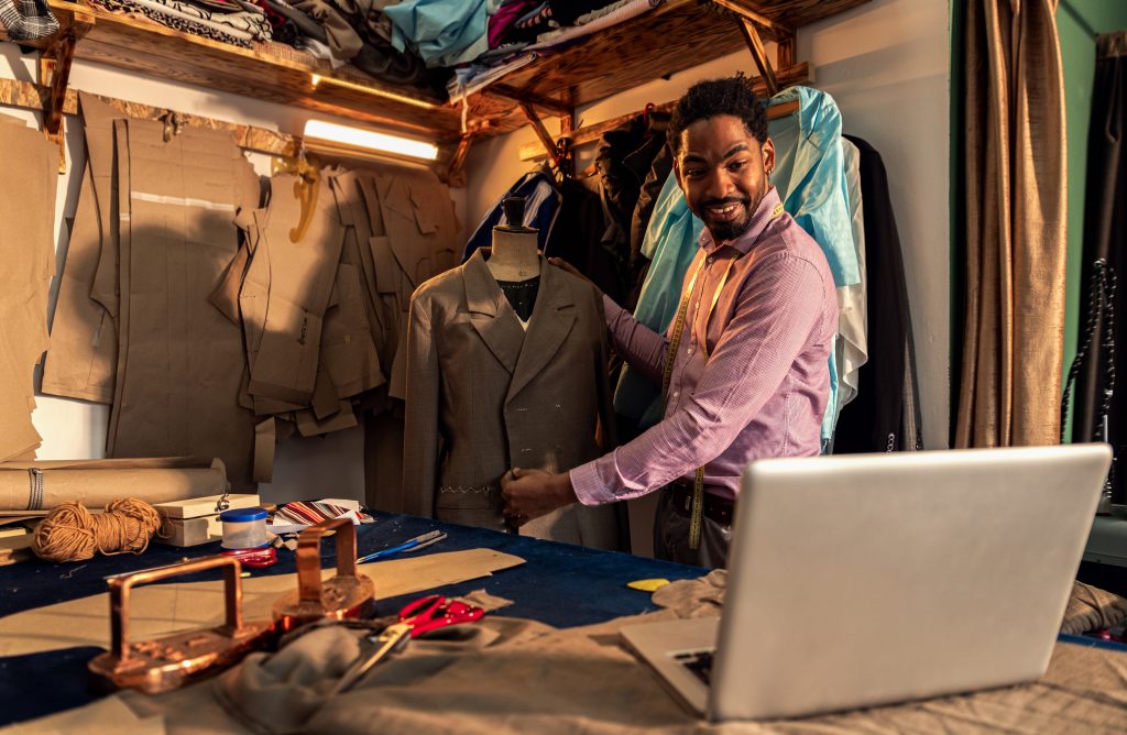 Tailor showcasing a suit jacket in a workshop while using a laptop, representing ABC Lifestyle Co. using an SMS Gateway Provider.