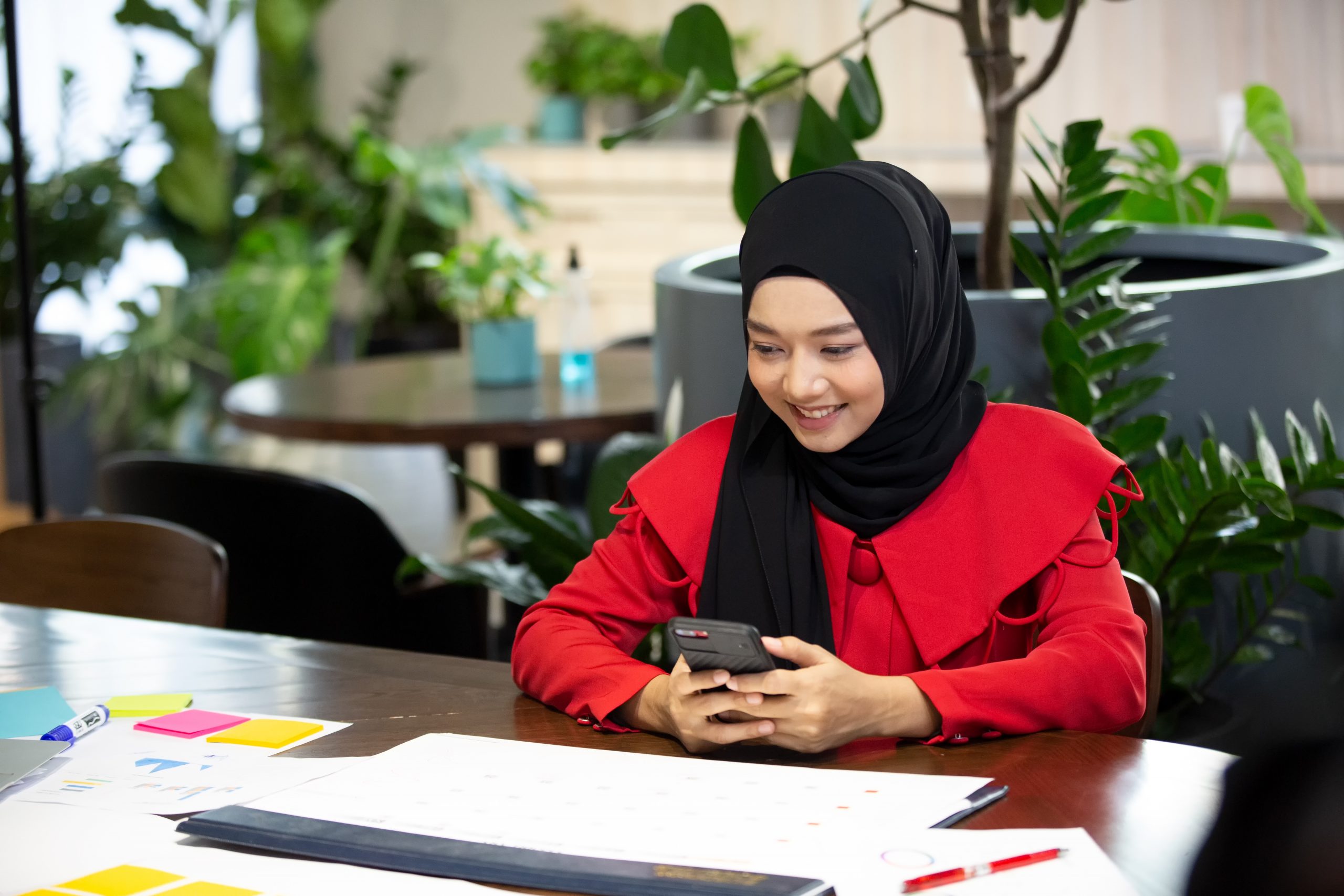 Muslim woman using her phone while working at a desk in a modern office, representing business use of SMS Provider Malaysia.