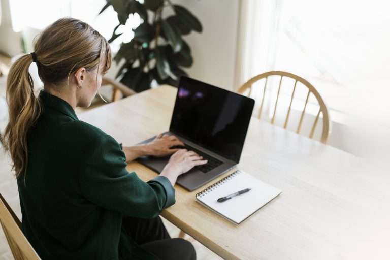 Woman working on a laptop at a wooden table with a notebook beside her, illustrating SaaS productivity using a programmable SMS API.