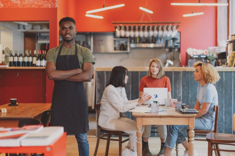 Restaurant owner standing confidently while customers dine inside a café, showing how sms marketing malaysia helps fill tables